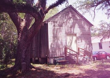 A white clapboard building sits beneath a canopy of trees. Stairs lead to a closed door on the front.