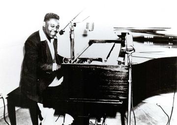 Fats Domino smiles at the camera from his piano, where he sits in front of a microphone