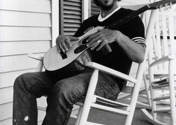 Bobby Charles sits on in a rocking chair holding a guitar on a home's front porch