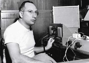 Cosimo, in a white T shirt and tweed pants, sits in front of recording equipment