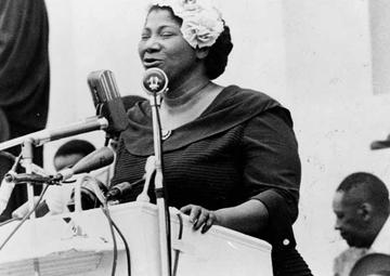 Mahalia Jackson sings from a podium on a stage. She wears a dark dress and has a fancy white hat on