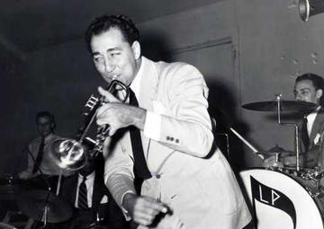 A trumpet-holding Louis Prima stands on stage with a band behind him. He wears a white suit with a black tie.