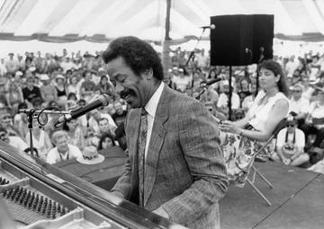 A black-and-white image of Allan playing the piano in front of a crowd gathered under an event tent