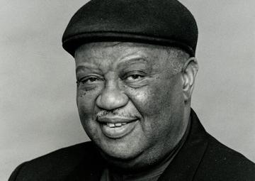 Portrait of Alvin Batiste wearing a dark hat and a jacket with a jaunty kerchief 