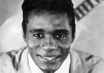 Eddie, in a sharp, light-colored suit and tie, stares into the camera. A guitar is on a backdrop behind him.