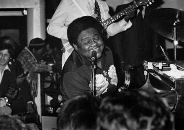 Fats sings into a microphone with a big smile on his face while playing the piano. A band plays behind him. 