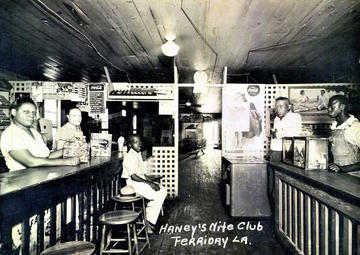 A group of people sit behind a counter top in a nightclub, all turned to face the camera. A younger person sits next to a straw hat. 