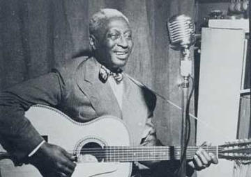 Leadbelly sits with his guitar in front of a microphone in a recording studio