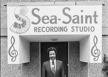 Allen, in a black suit and tie, stands in front of the entrance to Sea-Saint Recording Studio. The sign for the studio features a swirling piano keyboard and two large music notes