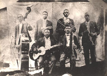 Buddy Bolder and his bandmates; four men stand with their instruments, and two sit; we see a base, guitars, trombones and clarinets