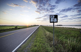 A rural two-lane highway, part of Louisiana's Cajun Corridor Byway, stretches into the distance. In the background the sun peeks over the horizon, brightening a cloudy sky.
