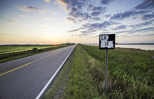 A rural two-lane highway, part of Louisiana's Cajun Corridor Byway, stretches into the distance. In the background the sun peeks over the horizon, brightening a cloudy sky.
