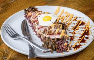 A teak has an egg cracked on top of it, drizzled with yellow and red sauces with a fork and knife to the side