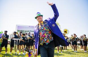 A person dressed in a festive blazer and hat waves to a crowd celebrating Mardi Gras.
