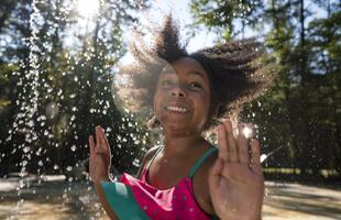 A young girl smiling while she plays in a splash pad, with water spraying around her.