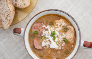 A white and brown mug of chicken and sausage gumbo, topped with white rice and sliced green onions, with slices of sausage and chunks of chicken visible through the well-seasoned broth.