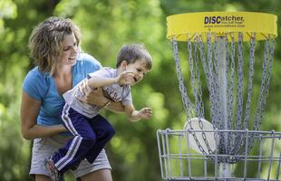 A mother holds her child, who reaches out to grab a disc from the disc golf basket.