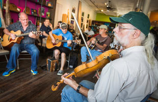 Four men sit in a circle, each holding a different instrument, including acoustic guitars and fiddles.