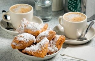 Two white mugs holding cafe au lait are set beside a shallow bowl of flaky beignets topped with powdered sugar from Cafe du Monde in New Orleans, Louisiana.